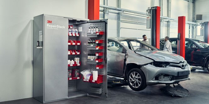 Two people talk next to a damaged vehicle and an open 3M™ RepairStack™ cabinet.
