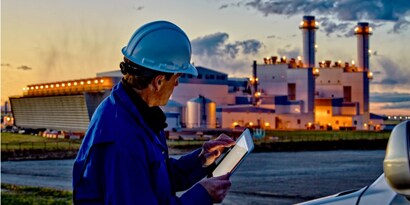 man in a construction hat on a tablet in front of an industrial plant
