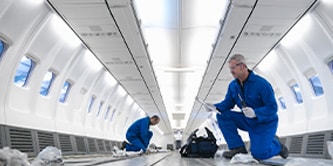 Two people in blue work gear and protective eyewear working on the inside of an airplane.
