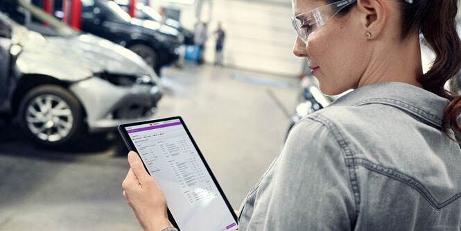 A woman standing in an autobody shop looks at the screen of a tablet device.

