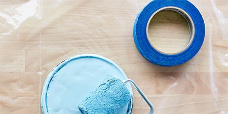Paint lid, paint roller & brush shown on top of a 3M masking film protecting a wood floor
