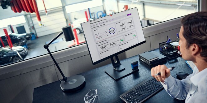 A man sitting in a room above an auto body shop looks at a dashboard on a desktop screen.
